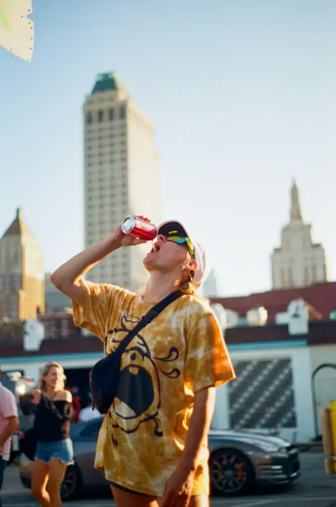 Woman in a yellow tie-dye shirt drinking from a can on a downtown street