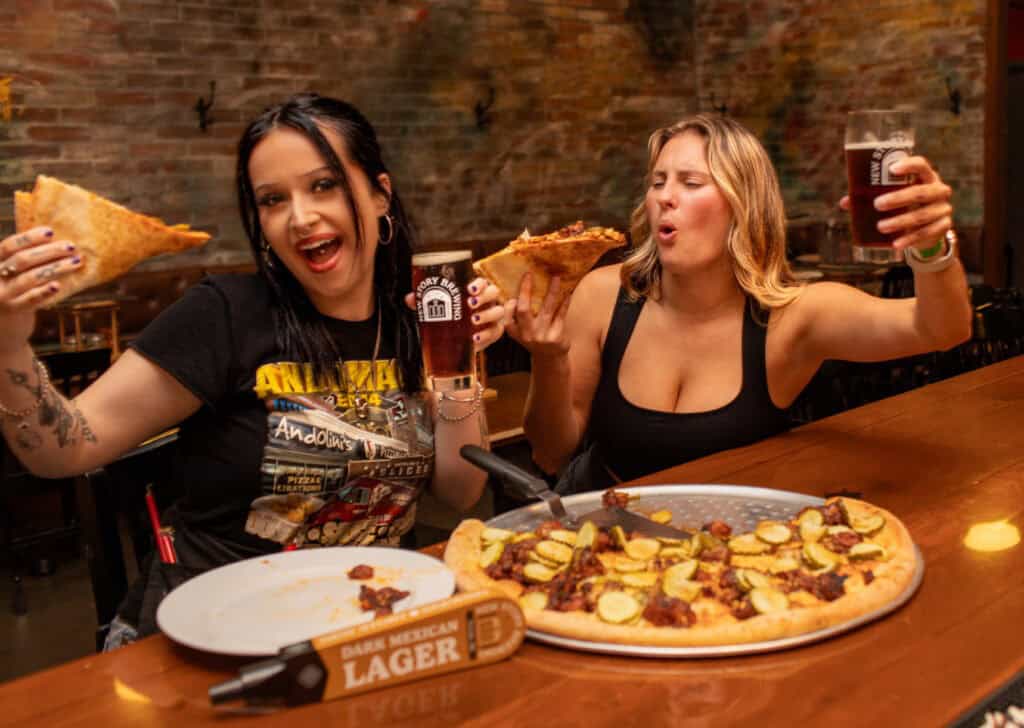 Two friends cheering with drinks over a pizza at a bar