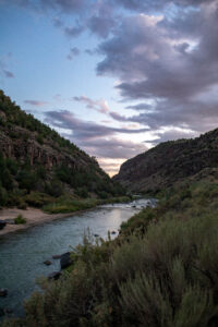 Scenic nature photograph of a wide river flowing through a sunlit canyon with lush green banks.