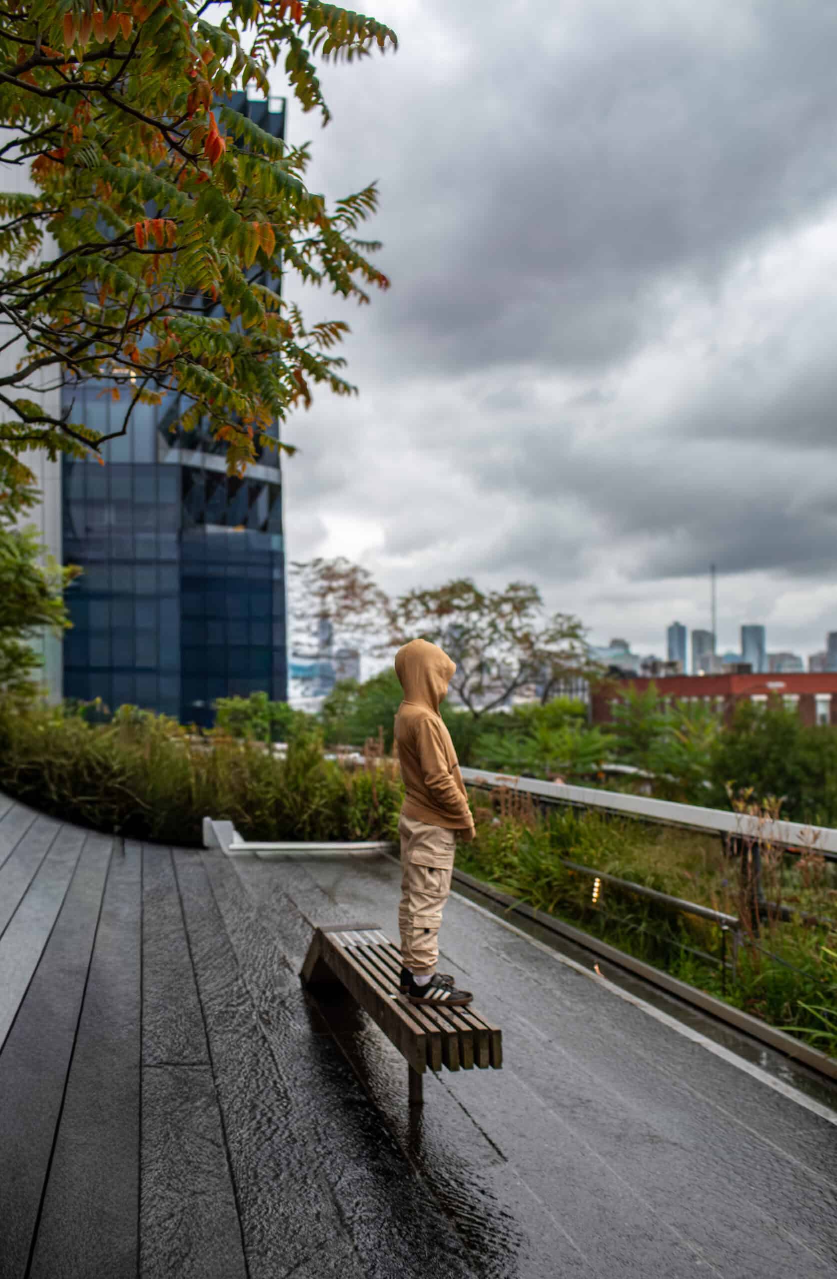 Person in hoodie standing on park bench with cityscape background.