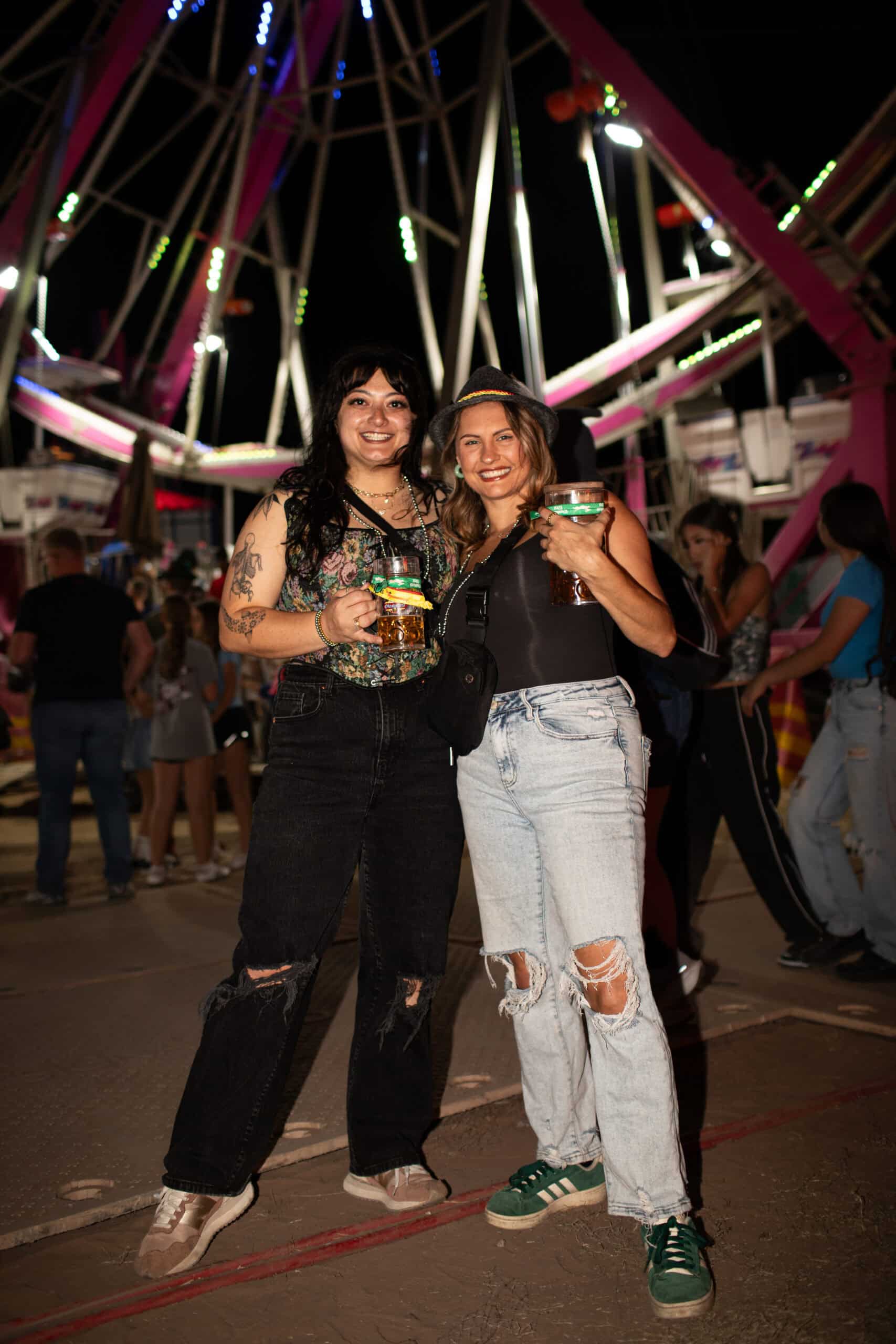Two women smiling at a nighttime fair, holding drinks, with a Ferris wheel in the background.