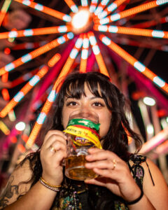 Woman smiling and eating at a night fair with a brightly lit Ferris wheel in the background, an example of event photography.