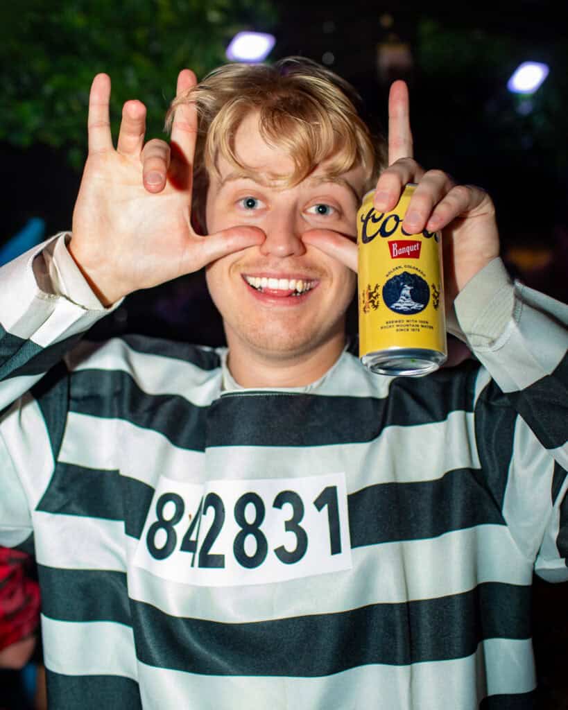Young man smiling and holding a beer can at a nighttime outdoor event.