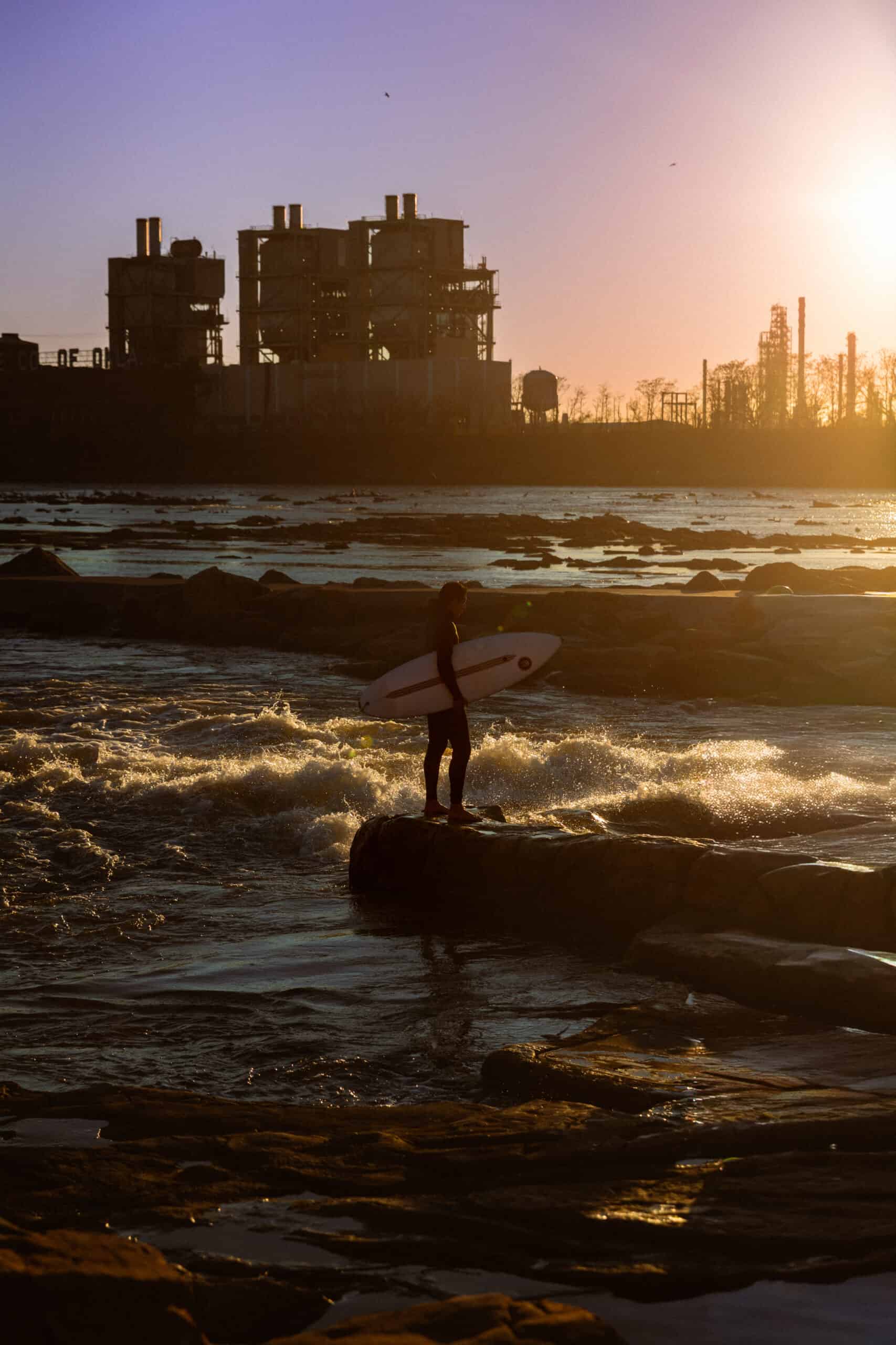 Surfer standing on a rock with a surfboard during sunset at the Oklahoma shoreline.