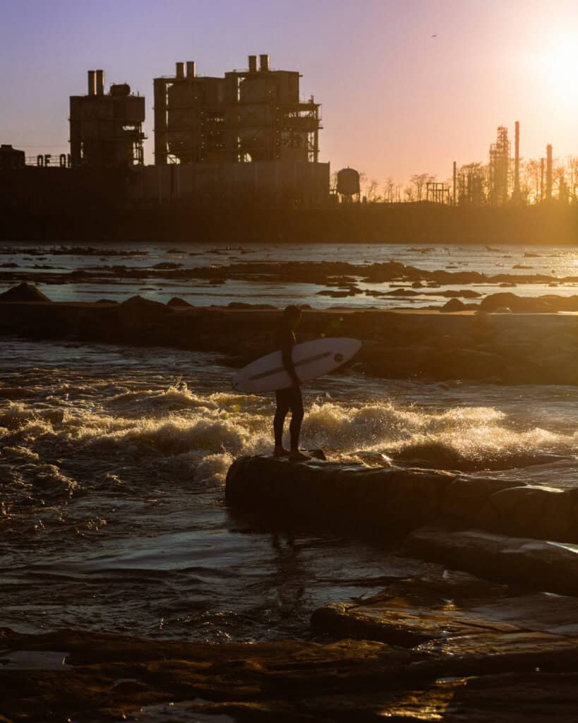 Surfer standing on rocks with surfboard at sunset near industrial area in Oklahoma.