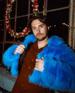 A young man poses confidently in a striking blue faux fur coat, indoors with holiday wreaths and war.