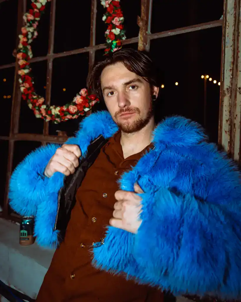 A young man poses confidently in a striking blue faux fur coat, indoors with holiday wreaths and war.