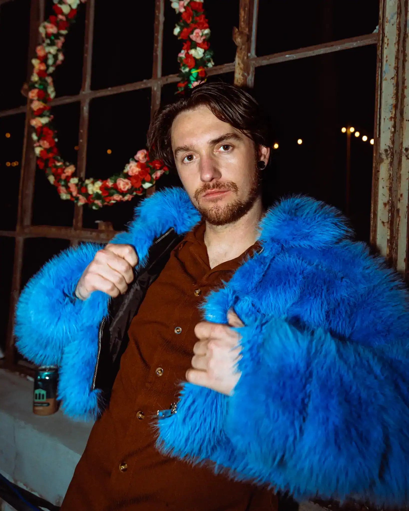 A young man poses confidently in a striking blue faux fur coat, indoors with holiday wreaths and war.