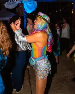 Rainbow-haired woman dancing at a lively party, wearing a colorful hat and shimmering outfit.
