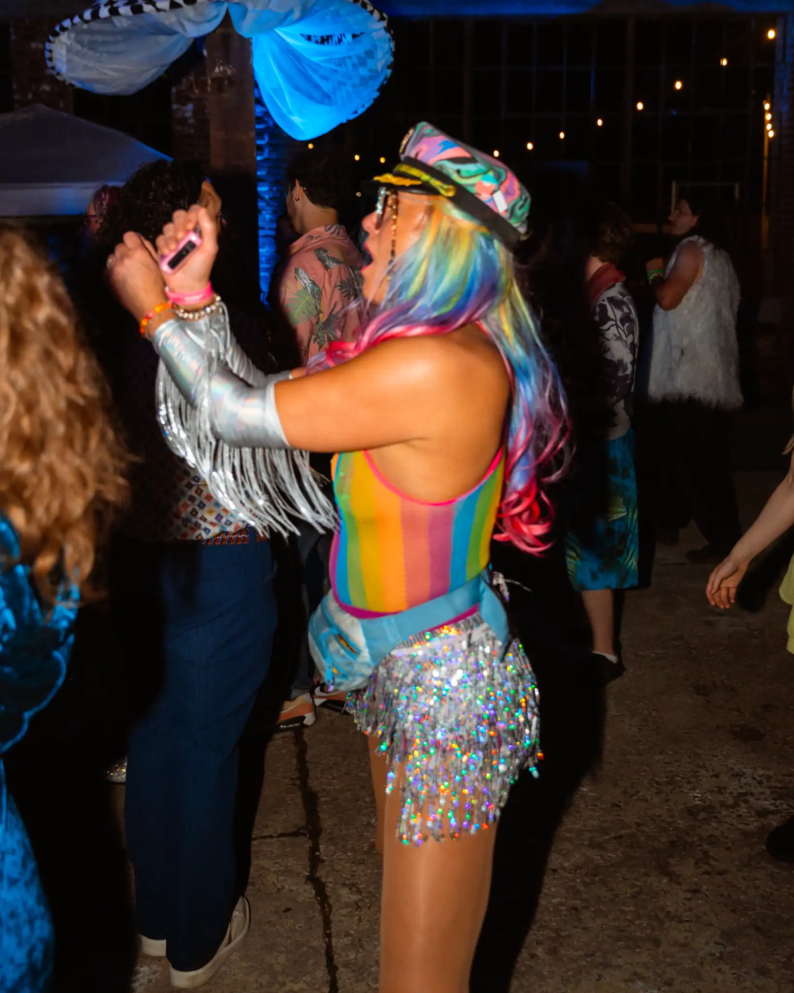 Rainbow-haired woman dancing at a lively party, wearing a colorful hat and shimmering outfit.