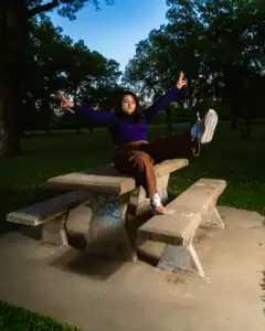 Young girl sitting on park bench with arms raised, enjoying outdoor scenery at sunset.