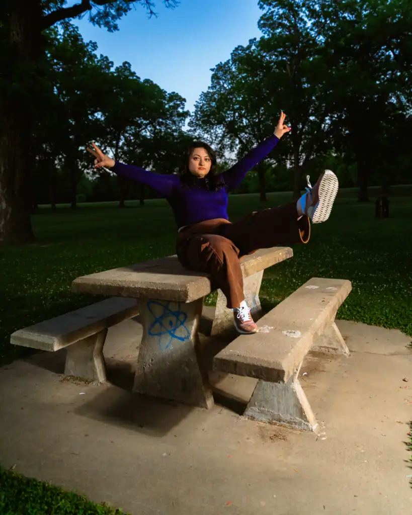 Young girl sitting on park bench with arms raised, enjoying outdoor scenery at sunset.