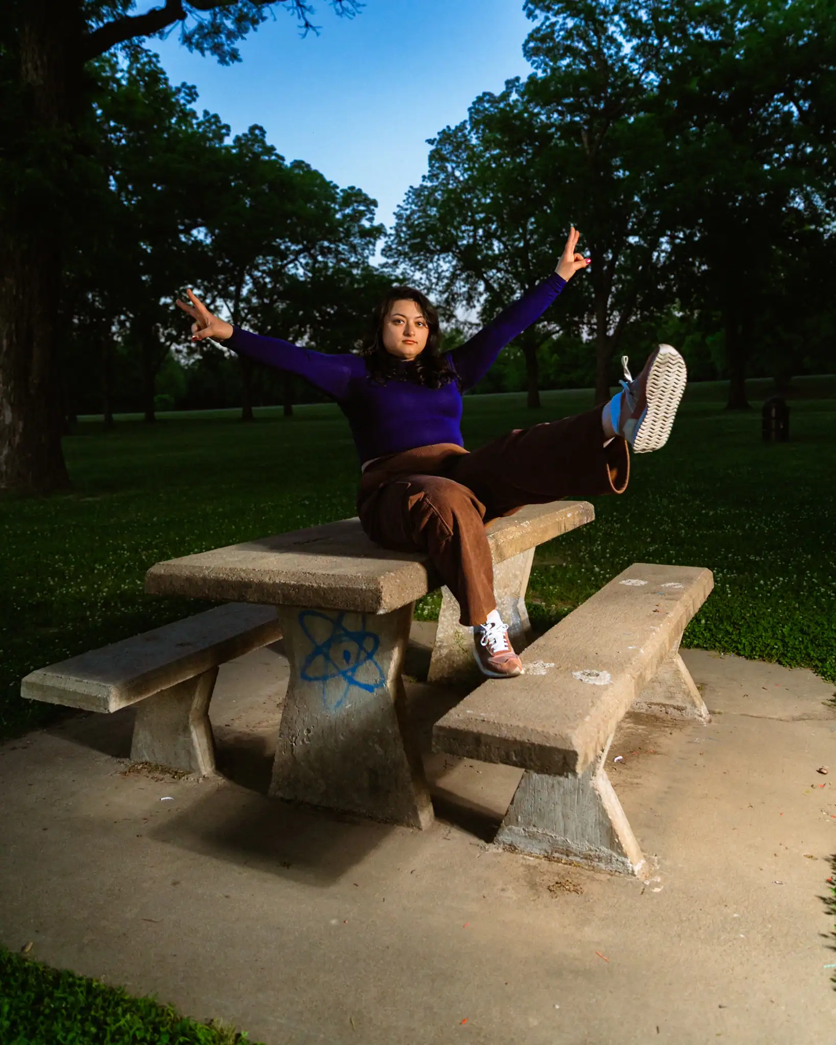 Young girl sitting on park bench with arms raised, enjoying outdoor scenery at sunset.