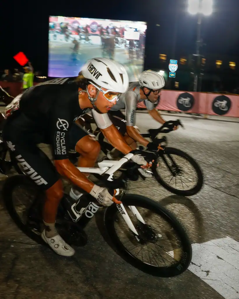Cyclists racing at night past a large video screen