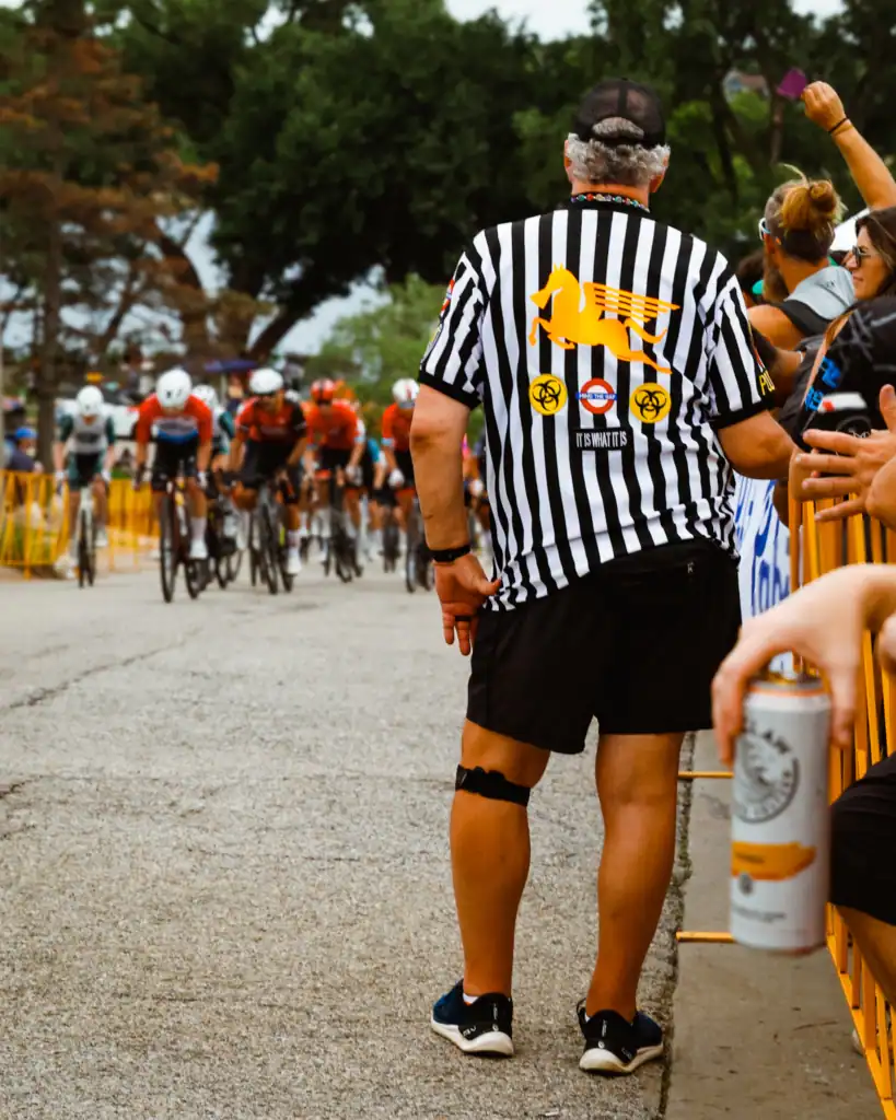 Race official in a striped shirt watching cyclists pass during a street race